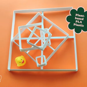 May include: White geometric cookie cutters in various sizes are arranged on an orange surface. A yellow rubber duck is also present. A green flower-shaped graphic reads "Plant-based PLA Plastic".
