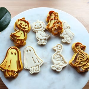 May include: A collection of Halloween-themed cookie cutters in the shape of ghosts, ducks, and jack-o'-lanterns. The cutters are made of yellow plastic and are arranged on a white marble surface. The cookies are in various stages of completion.