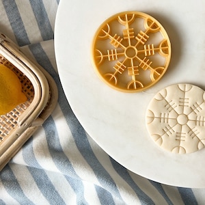 May include: Two cookie cutters in the shape of a snowflake with a geometric design. The cutter on the left is gold and the cutter on the right is white.