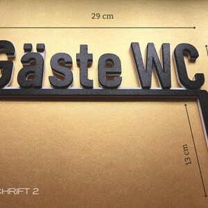 May include: Black and white sign that reads "G&auml;ste WC". The sign is 29 cm long and 8 cm high, with an additional 13 cm long section. The text is in a bold, 3D font. The sign is on a gold background.