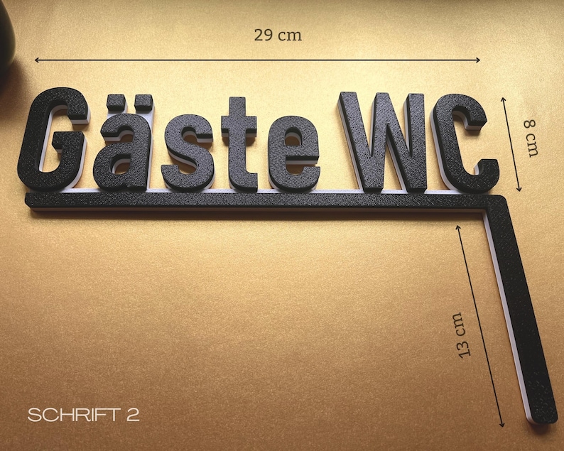 May include: Black and white sign that reads "G&auml;ste WC". The sign is 29 cm long and 8 cm high, with an additional 13 cm long section. The text is in a bold, 3D font. The sign is on a gold background.