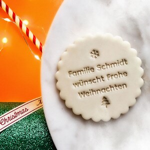 May include: White round cookie with a scalloped edge, decorated with a snowflake and a Christmas tree. The cookie has the text "Familie Schmidt w&uuml;nscht Frohe Weihnachten" in brown.