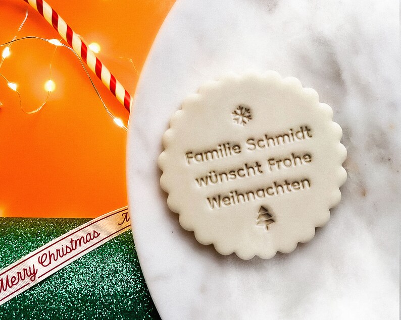 May include: White round cookie with a scalloped edge, decorated with a snowflake and a Christmas tree. The cookie has the text "Familie Schmidt w&uuml;nscht Frohe Weihnachten" in brown.