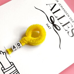 以下が含まれることがあります： ドイツ語の「ALLES」という単語と「F&Uuml;R JEMANDEN DER SCHON HAT」というフレーズが黒い文字で書かれた白いカード。白いビーズに「40」の数字が入った黄色のふわふわリングがカードに付いています。漫画の手も表示されています。