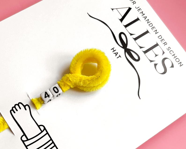 以下が含まれることがあります： ドイツ語の「ALLES」という単語と「F&Uuml;R JEMANDEN DER SCHON HAT」というフレーズが黒い文字で書かれた白いカード。白いビーズに「40」の数字が入った黄色のふわふわリングがカードに付いています。漫画の手も表示されています。