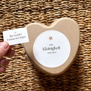 May include: A heart-shaped, light brown gift box with a white circular label that reads "Eine Kleinigkeit f&uuml;r Euch." A small card held nearby says "Ihr werdet Uroma & Uropa."