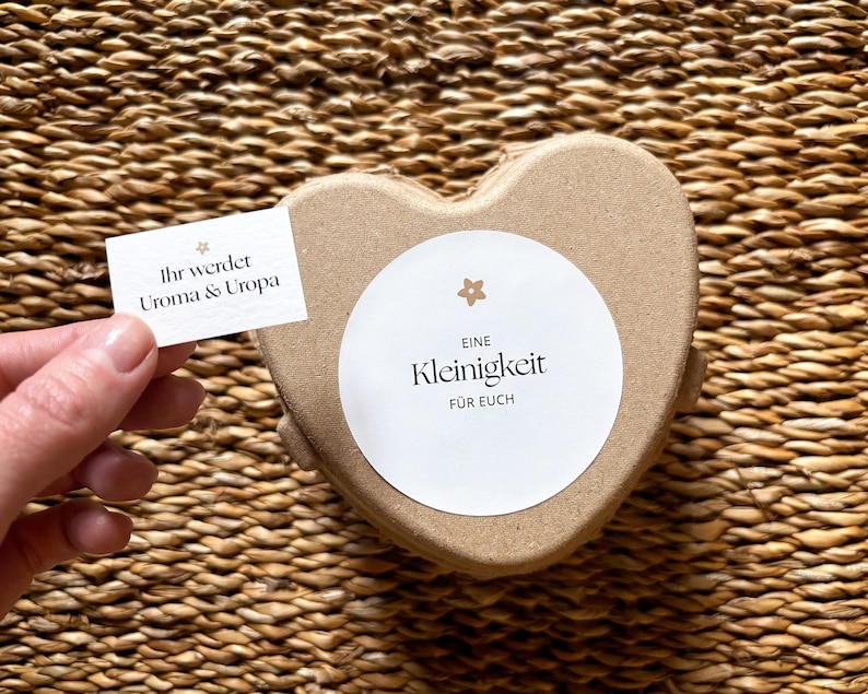 May include: A heart-shaped, light brown gift box with a white circular label that reads "Eine Kleinigkeit f&uuml;r Euch." A small card held nearby says "Ihr werdet Uroma & Uropa."