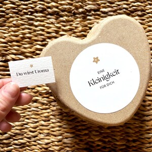 May include: A heart-shaped, light brown box with a white circular label that reads "Eine Kleinigkeit f&uuml;r Dich". A small card held by a hand reads "Du wirst Uroma". The background is a woven texture.