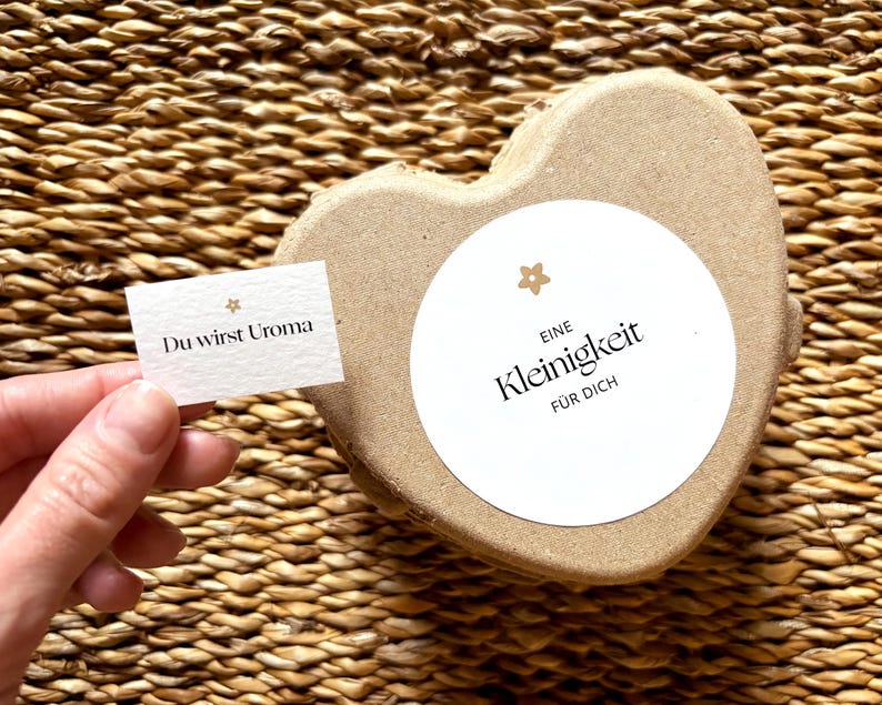 May include: A heart-shaped, light brown box with a white circular label that reads "Eine Kleinigkeit f&uuml;r Dich". A small card held by a hand reads "Du wirst Uroma". The background is a woven texture.
