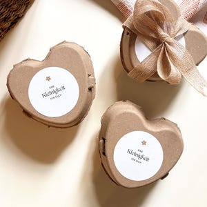 May include: Three heart-shaped, brown cardboard boxes with white circular labels. The labels have the text "Eine Kleinigkeit f&uuml;r dich" and a small star. One box is tied with a brown ribbon. The boxes are on a light-colored surface.