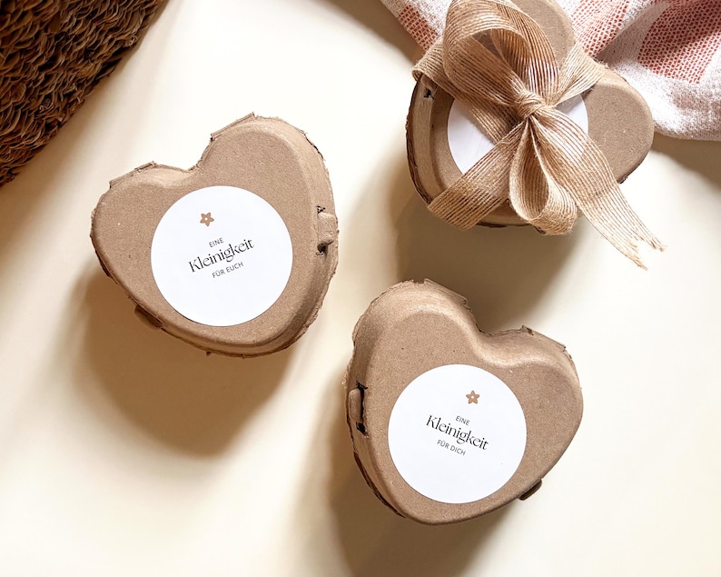 May include: Three heart-shaped, brown cardboard boxes with white circular labels. The labels have the text "Eine Kleinigkeit f&uuml;r dich" and a small star. One box is tied with a brown ribbon. The boxes are on a light-colored surface.