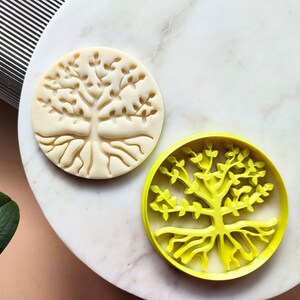 May include: Two cookie cutters in the shape of a tree with roots and branches. One cutter is white and the other is yellow.