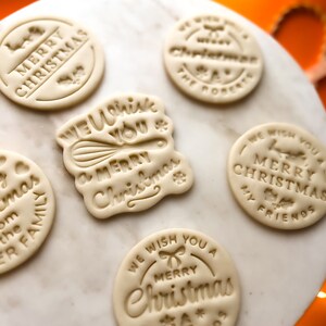 May include: Five round, white, cookie dough ornaments with different Christmas greetings. The greetings include "Merry Christmas", "We Wish You a Merry Christmas", "We Wish You a Merry Christmas My Friends", and "Merry Christmas from the Miller Family".