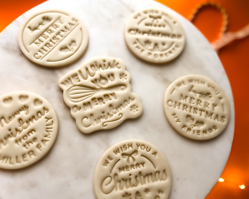 May include: Five round, white, cookie dough ornaments with different Christmas greetings. The greetings include "Merry Christmas", "We Wish You a Merry Christmas", "We Wish You a Merry Christmas My Friends", and "Merry Christmas from the Miller Family".