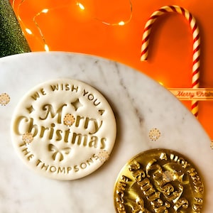 May include: A festive Christmas-themed image featuring a white cookie stamp with the words "We Wish You a Merry Christmas" and "The Thompsons" engraved. A gold-colored stamp is also visible. A candy cane and decorative lights complete the holiday scene.