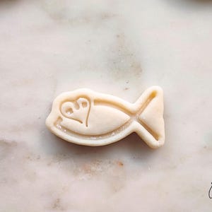 May include: A light-colored fish-shaped cookie cutter with a heart and foot design. The cookie cutter is on a marble surface. The cookie cutter has a tail and fins. The cookie cutter is a baking tool.