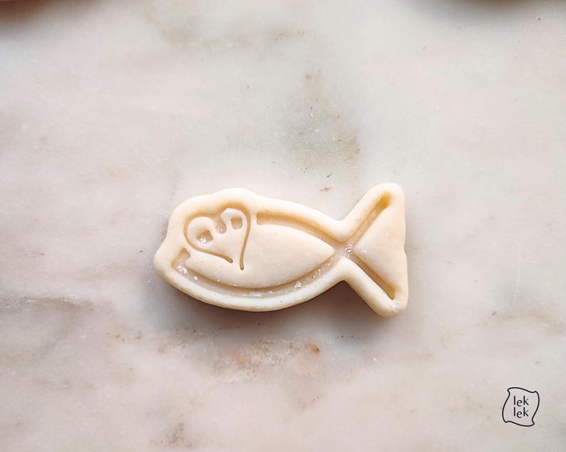 May include: A light-colored fish-shaped cookie cutter with a heart and foot design. The cookie cutter is on a marble surface. The cookie cutter has a tail and fins. The cookie cutter is a baking tool.