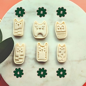 May include: Six cat-shaped cookie cutters arranged on a white marble surface. Each cutter has a unique cat face design. Green flower-shaped stickers with numbers 1-6 are placed around the cutters. The background is a mix of orange and green.