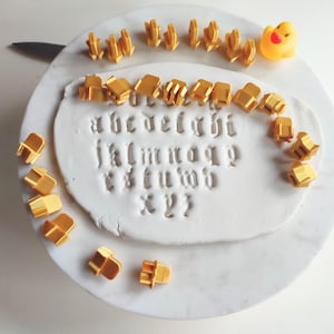 Puede incluir: Una superficie de arcilla blanca con el alfabeto estampado, rodeada de cortadores de letras doradas. Un patito de goma amarillo está en el borde. Un cuchillo es visible en el lado izquierdo. Las letras están en fuente gótica.