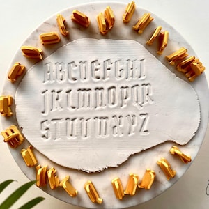May include: A set of gold-colored plastic alphabet stamps arranged in a circle around a white clay slab. The clay slab features the full English alphabet from A to Z embossed in a gothic-style font. The stamps are individual pieces for each letter.