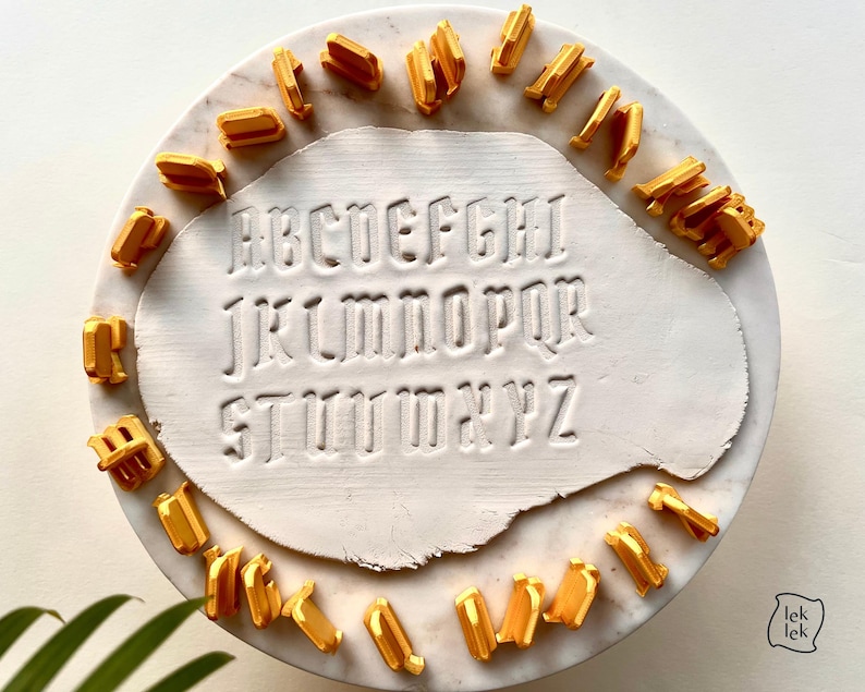 May include: A set of gold-colored plastic alphabet stamps arranged in a circle around a white clay slab. The clay slab features the full English alphabet from A to Z embossed in a gothic-style font. The stamps are individual pieces for each letter.