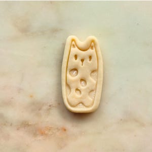 May include: A light-colored cat-shaped cookie cutter with a rectangular outline. The cat's face and body are detailed with cut-out shapes. The cutter is made of a light-colored material and is set against a marble background.