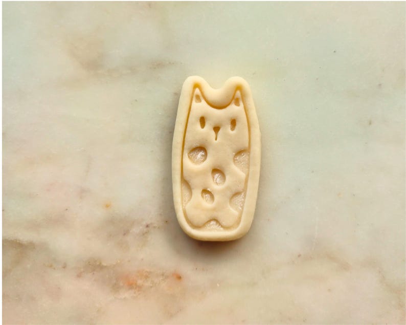 May include: A light-colored cat-shaped cookie cutter with a rectangular outline. The cat's face and body are detailed with cut-out shapes. The cutter is made of a light-colored material and is set against a marble background.