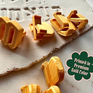 May include: A collection of three-dimensional gold-colored letters with a textured surface finish. A dark green scalloped badge features the white text Printed in Premium Gold Color. The letters are arranged on a light-colored surface.
