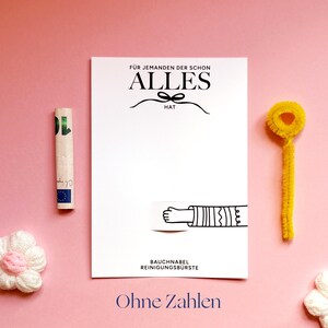 May include: A white rectangular card with black text reading F&Uuml;R JEMANDEN DER SCHON ALLES HAT and BAUCHNABEL REINIGUNGSB&Uuml;RSTE, featuring a line drawing of an arm. Beside it are a rolled ten euro bill, a yellow pipe cleaner, and two white flowers.