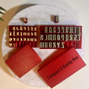 May include: A set of gold-colored metal alphabet stamps arranged in two red plastic trays. The set includes uppercase and lowercase letters. Two red rectangular storage boxes are included, one of which features the text Compact & Handy Box.