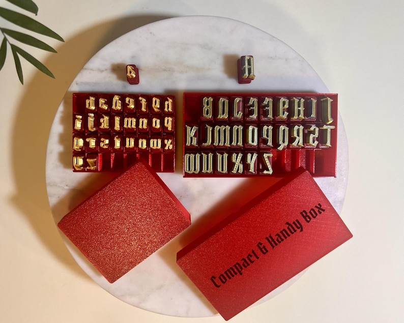 May include: A set of gold-colored metal alphabet stamps arranged in two red plastic trays. The set includes uppercase and lowercase letters. Two red rectangular storage boxes are included, one of which features the text Compact & Handy Box.