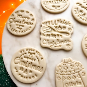 May include: Assortment of six white sugar cookies decorated with Christmas-themed designs. The cookies feature phrases like "We Wish You a Merry Christmas", "Merry Christmas from the Miller Family", and "Merry Christmas My Friends".