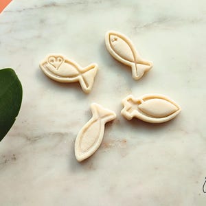 May include: Four beige fish-shaped cookie cutters on a marble surface. One cutter has a heart design, while the others have simple fish outlines. The cutters are arranged diagonally, with a green leaf and a logo in the corner.