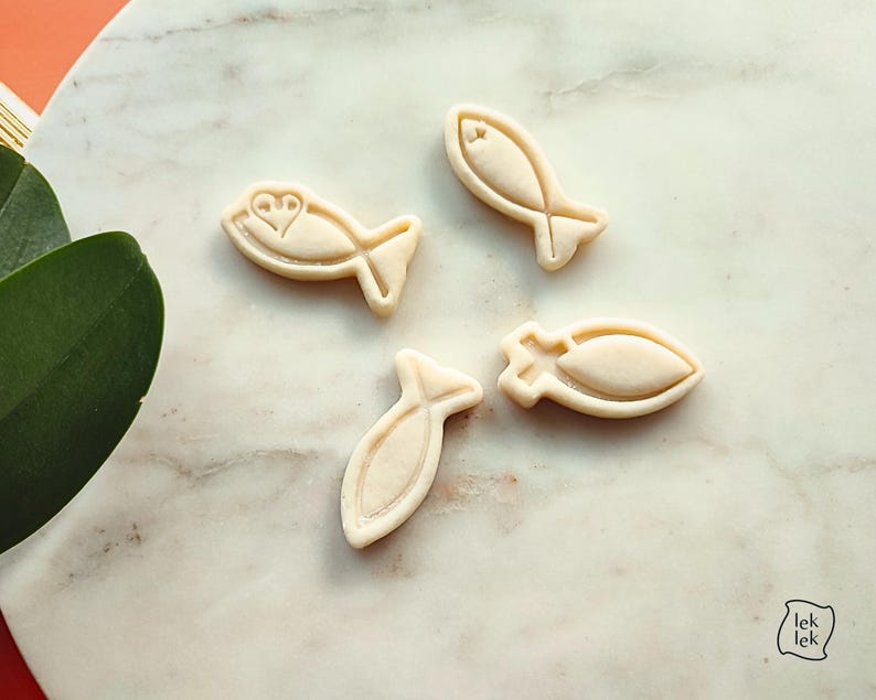 May include: Four beige fish-shaped cookie cutters on a marble surface. One cutter has a heart design, while the others have simple fish outlines. The cutters are arranged diagonally, with a green leaf and a logo in the corner.