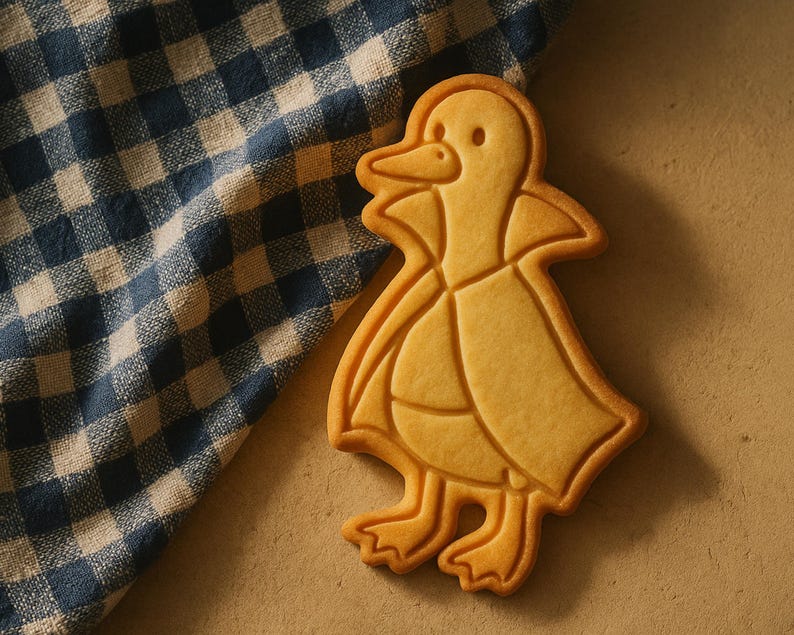 May include: A golden-brown duck-shaped cookie with detailed outlines, resting on a beige surface next to a blue and white checkered cloth. The cookie has a detailed beak, eyes, and feet.