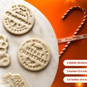 May include: Four white cookie dough rounds with Christmas-themed designs, including "We Wish You A Merry Christmas The Roberts", "We Wish You A Merry Christmas My Friends", "Think You Merry Christmas", and a snowflake design. A red and white candy cane and a white ribbon with the text "Merry Christmas" are in the background. The text "2 SIZES AVAILABLE - 3 inches (7.6 cm) 3.5 inches (8.9 cm)" is also visible.