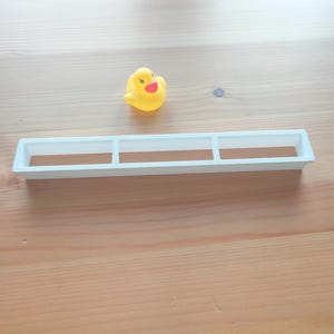 Puede incluir: Un patito de goma amarillo con pico rojo y ojos negros est&aacute; cerca de una bandeja rectangular blanca con tres compartimentos. Los objetos est&aacute;n sobre una superficie de madera marr&oacute;n claro.