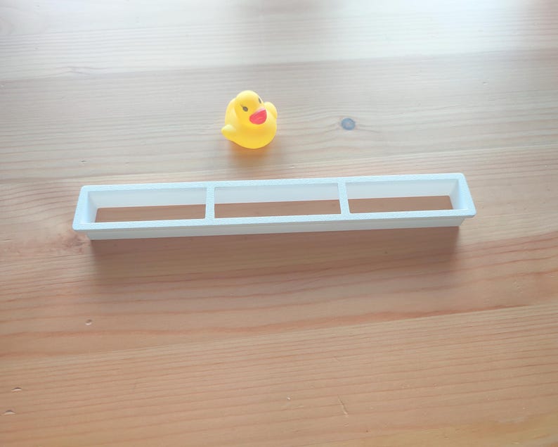 Puede incluir: Un patito de goma amarillo con pico rojo y ojos negros est&aacute; cerca de una bandeja rectangular blanca con tres compartimentos. Los objetos est&aacute;n sobre una superficie de madera marr&oacute;n claro.
