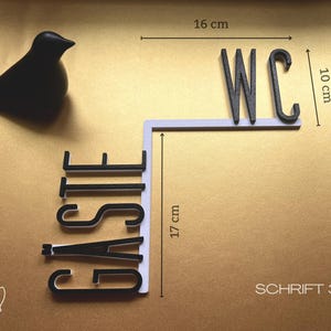 May include: A black decorative bird figurine and a black and white sign that reads "WC" (16 cm wide, 10 cm high) and "GASTE" (17 cm high). The sign is on a gold background. The text "SCHRIFT 3" is also visible.