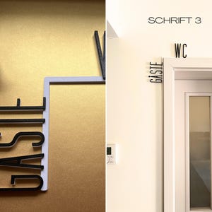 May include: Black and white decorative wall signs. One sign reads "G&auml;ste" in a bold, sans-serif font, with a black bird figurine. Another sign reads "WC" above a white door, with "G&auml;ste" on the side. The text "SCHRIFT 3" is above the door.