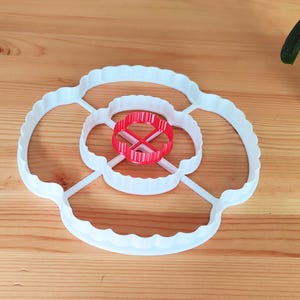 May include: White plastic flower-shaped cookie cutter with a smaller red plastic circle cutter inside.