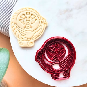 Könnte beinhalten: Zwei Ausstecher, einer rot und einer weiß, mit einem stilisierten Design eines Vogels mit ausgebreiteten Flügeln und einem Symbol in der Mitte. Das Symbol ist ein stilisiertes Kreuz mit einem Kreis darum.