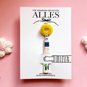 May include: A white rectangular card featuring the text F&Uuml;R JEMANDEN DER SCHON ALLES HAT and BAUCHNABEL REINIGUNGSB&Uuml;RSTE. A yellow pipe cleaner circle sits above a rolled banknote held by a printed illustration of an arm, with two white beads showing 30.
