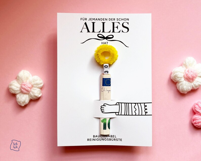 May include: A white rectangular card featuring the text F&Uuml;R JEMANDEN DER SCHON ALLES HAT and BAUCHNABEL REINIGUNGSB&Uuml;RSTE. A yellow pipe cleaner circle sits above a rolled banknote held by a printed illustration of an arm, with two white beads showing 30.