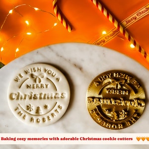 May include: Two white and gold Christmas cookie cutters. The white cutter says "We wish you a Merry Christmas The Roberts". The gold cutter says "A Joy to the World".