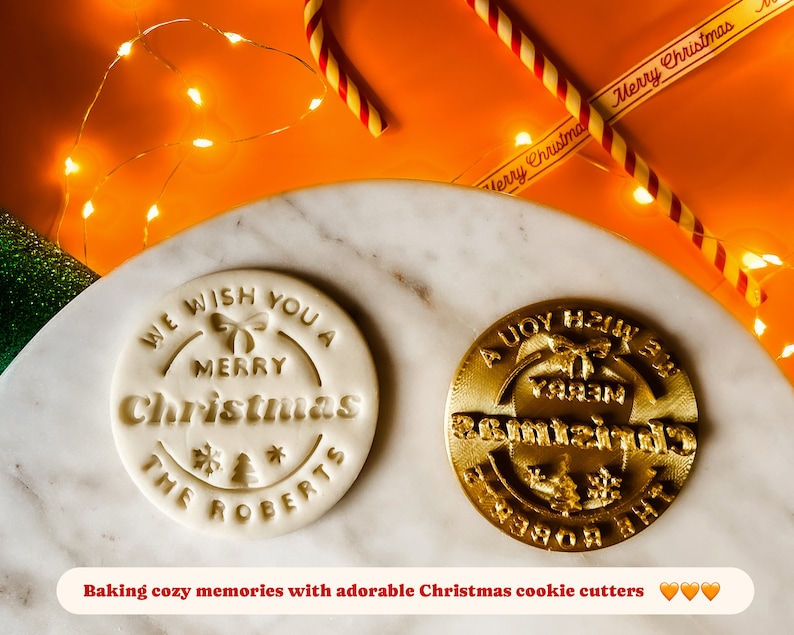 May include: Two white and gold Christmas cookie cutters. The white cutter says "We wish you a Merry Christmas The Roberts". The gold cutter says "A Joy to the World".