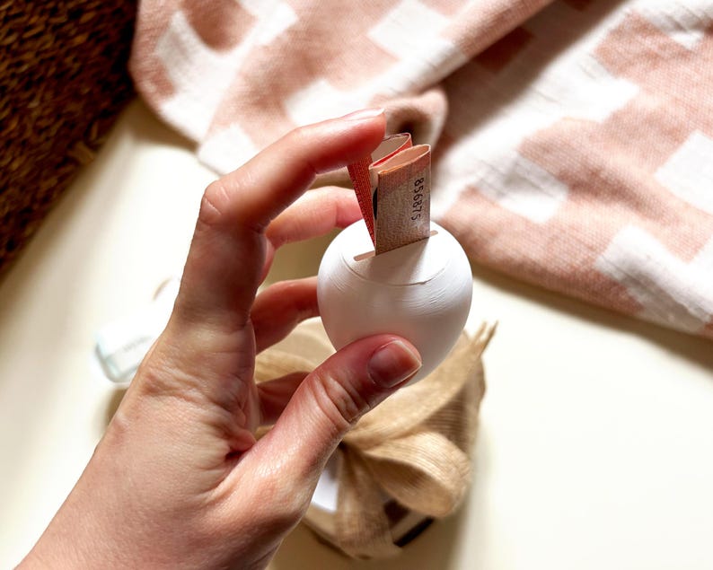 Pu&ograve; includere: Un oggetto in ceramica bianca a forma di uovo con una fessura, tenuto in mano. Una piccola pila di carta viene inserita nella fessura. Un tessuto a fantasia beige e bianco &egrave; sullo sfondo.