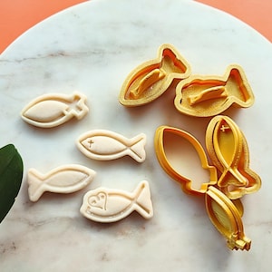 Puede incluir: Un surtido de galletas beige con forma de pez y cortadores de galletas dorados. Las galletas tienen detalles de cruz y corazón. Los cortadores tienen forma de pez, con un diseño interior detallado.