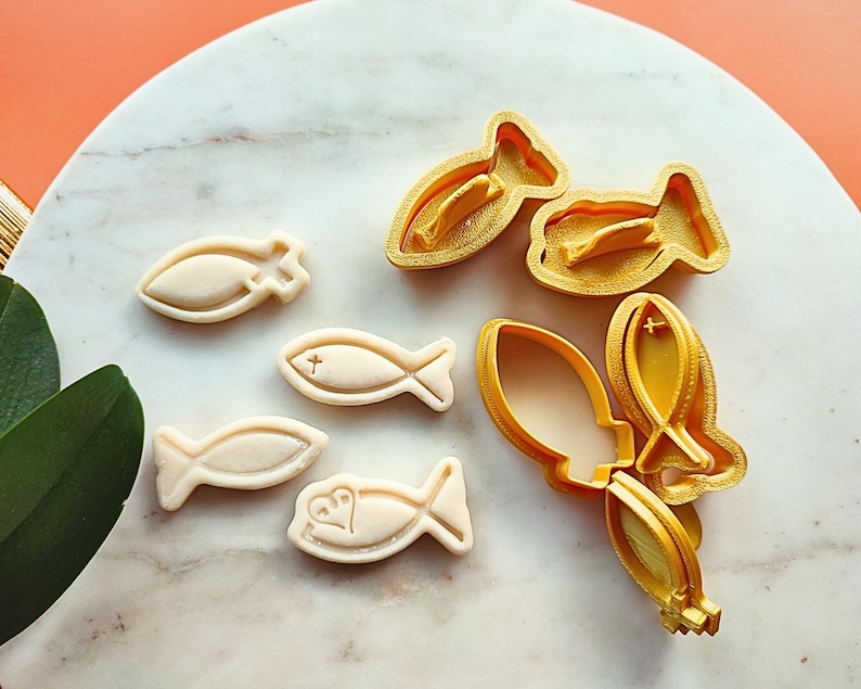 May include: A collection of fish-shaped cookie cutters and cookies. The cookies are off-white and feature cross and heart designs. The cutters are gold-colored plastic. The items are arranged on a white and marble surface.