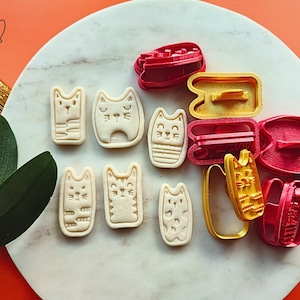 May include: A collection of cat-shaped cookie cutters and stamped cookies. The cutters are red and yellow, while the cookies are beige. The image is on a white marble surface with a bright orange background. The logo "lek lek" is in the top left corner.